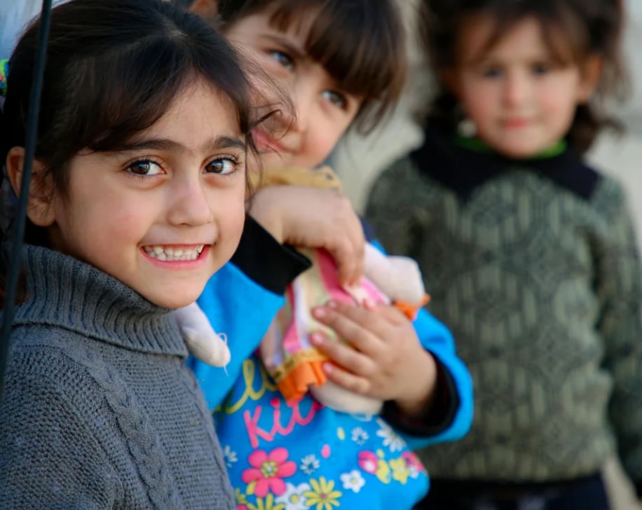 Three young children standing close together outdoors, the nearest smiling towards the camera.