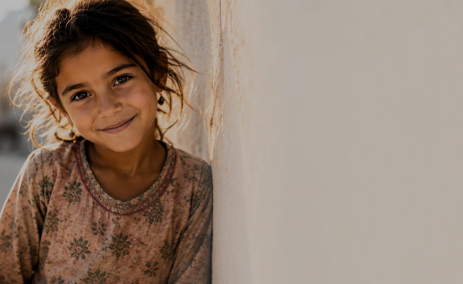 A girl leaning against a sunlit wall, smiling towards the camera.