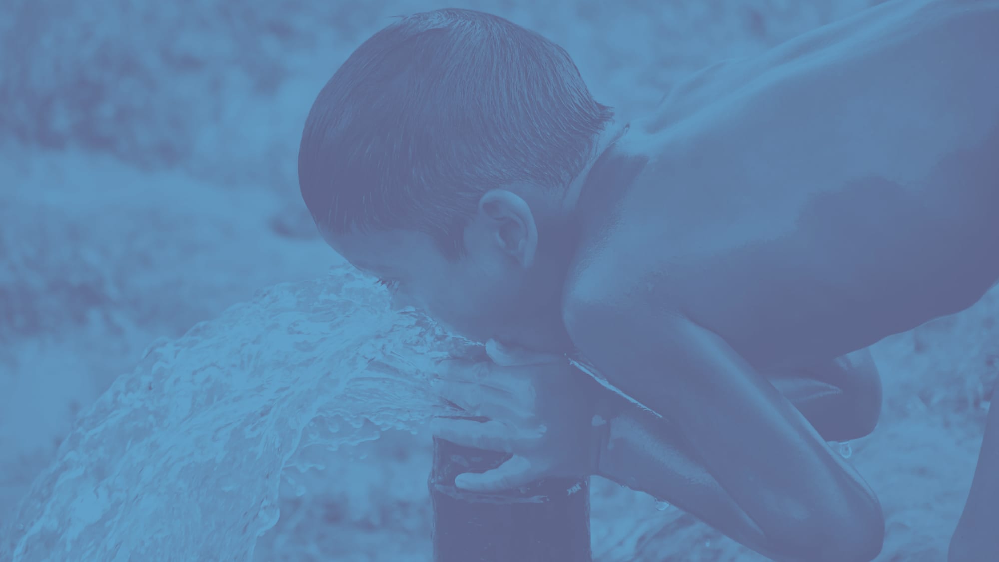 A boy drinking from the jet of a hand pump, cupping the water to his mouth.