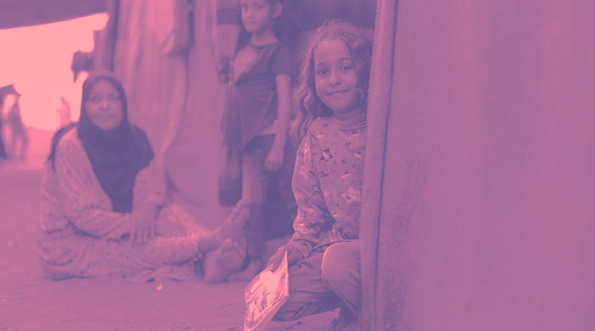 A girl sitting in the doorway of a tent shelter, her family gathered behind her.