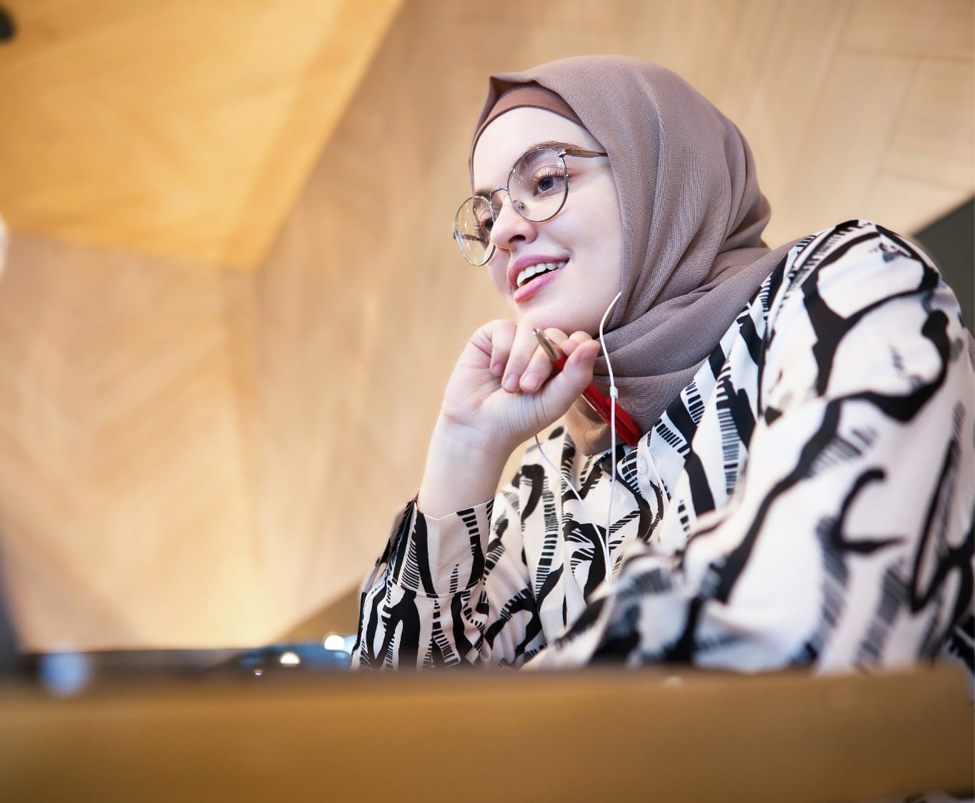 A young woman in glasses and a headscarf smiling, her chin resting on her hands.